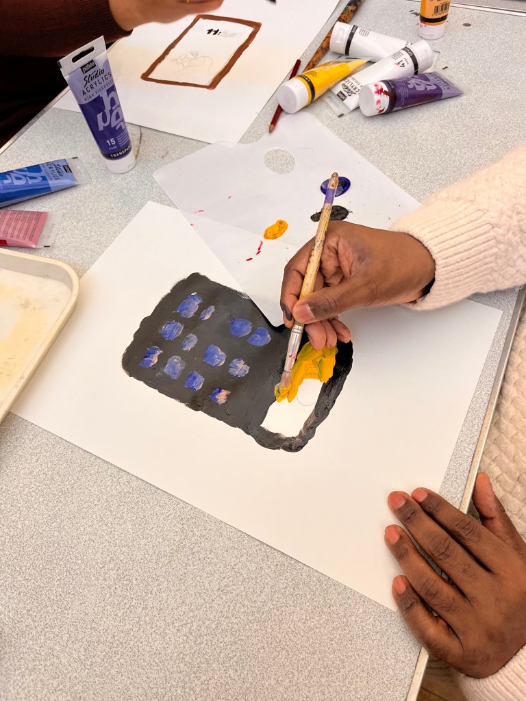 photo of people creating work. Two people are working on thei objects. One is a red rectangle. The other is a black square with blue buttons and yellow screen.