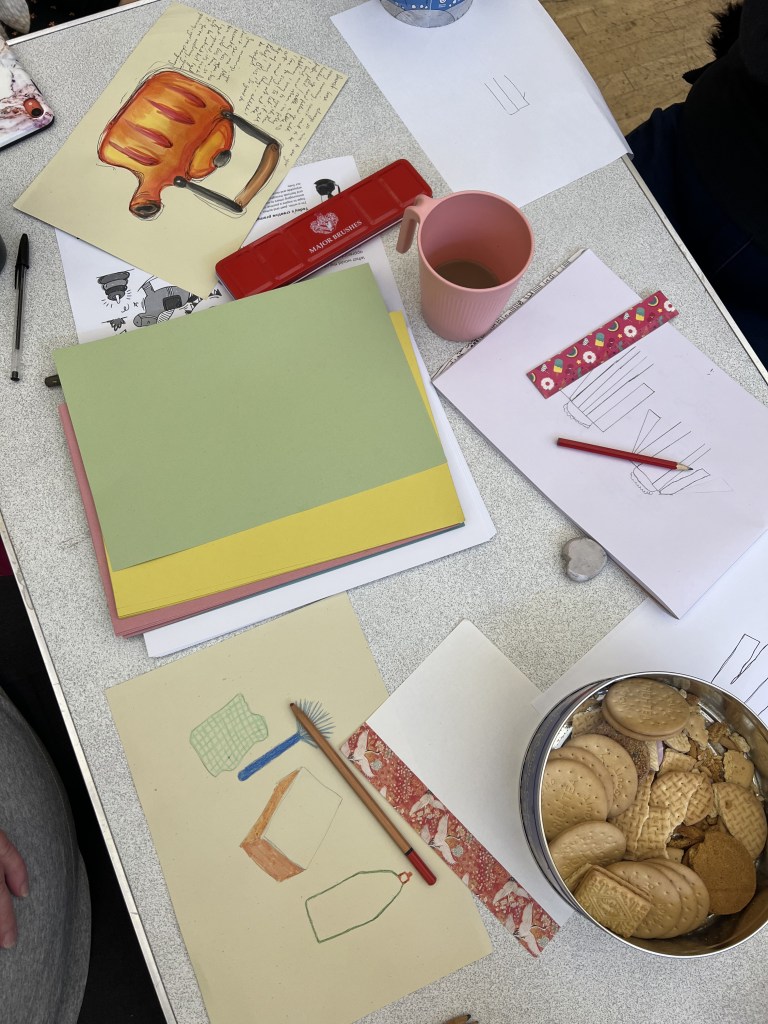 Various pieces of artwork on a table. one sheet has a orange kettle with writing next to it are some doodles and then theres another sheet with various objects such as sponges, bottles and duster. they are surrounded by paper, at supplies and theres a barrel of biscuits in the corner.