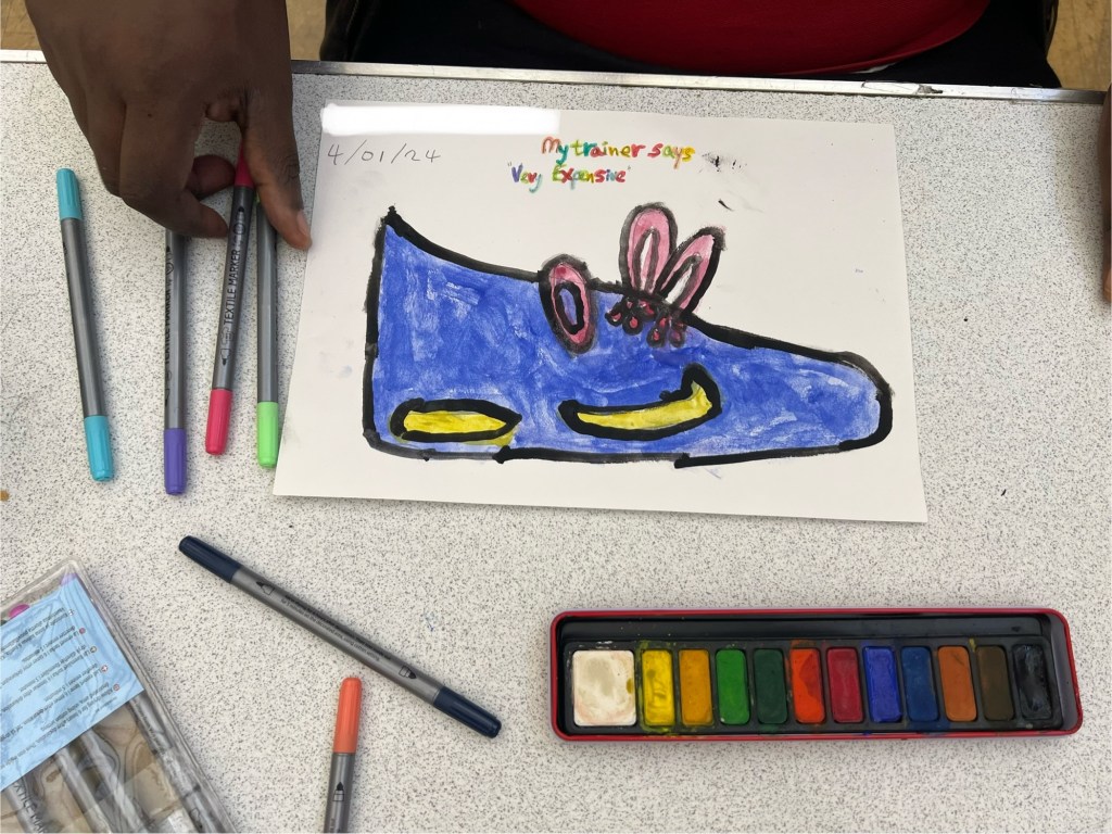 photoof artwork, painted with Gouache. Its a ultramarine blue trainer with red laces and yellow ticks below the laces. the arwork is on a table surrounded by coloured pens and a Gouache pan.
