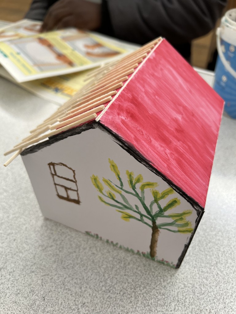 model of house. it is the exterior. The roof is red with one side using matches as hatching. on the wall is a window drawn in brown and on the side is a tree with yellow flowers on it.