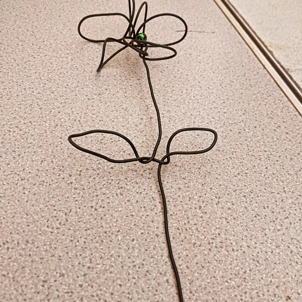 Example of wire work. Its shaped into a flower with a green bead for the center. The stem is long and extends beyond the photo.