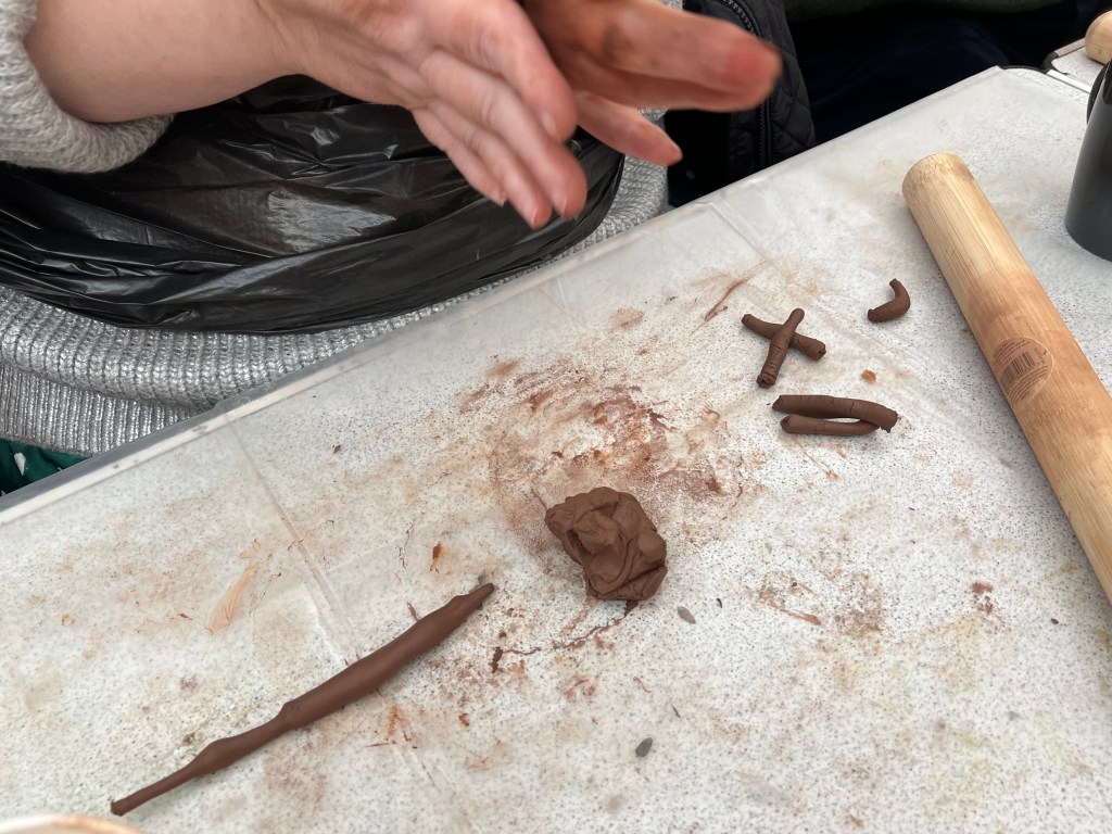 photo of someone rolling clay into a ball. On the table below participants hands is some clay shaped into a cross and a coil.