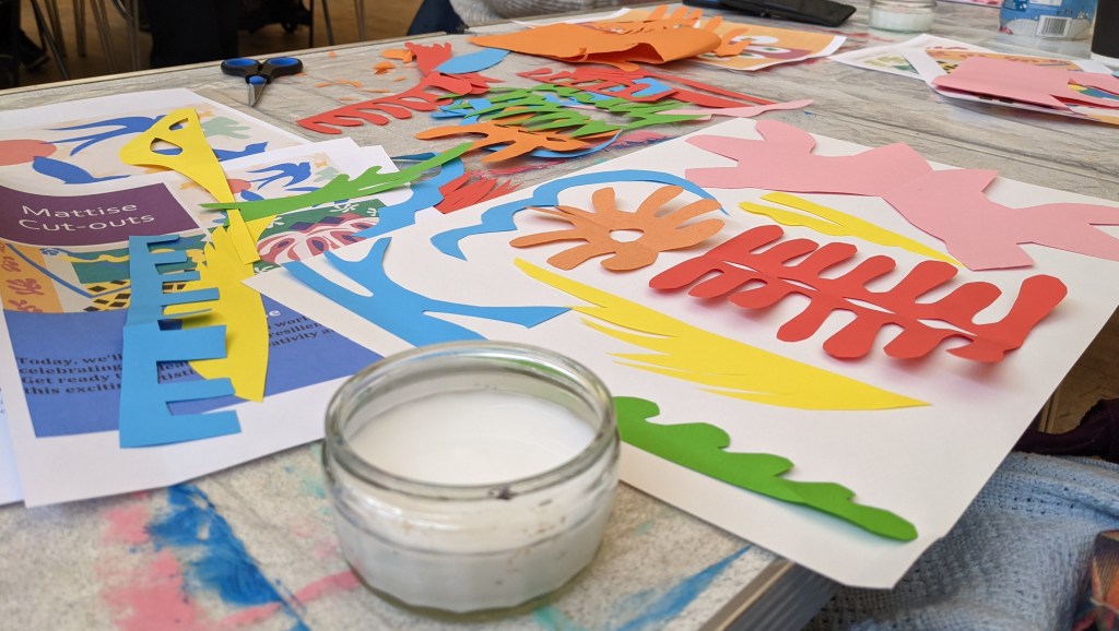 Photo of someone arranging a pile of shapes they have cut out- there are mutliple colours such as orange, blue, red and green.