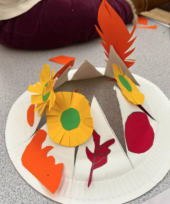 Halloween hat made out of paper plates. Has various yellow flowers and orange leaves attached.