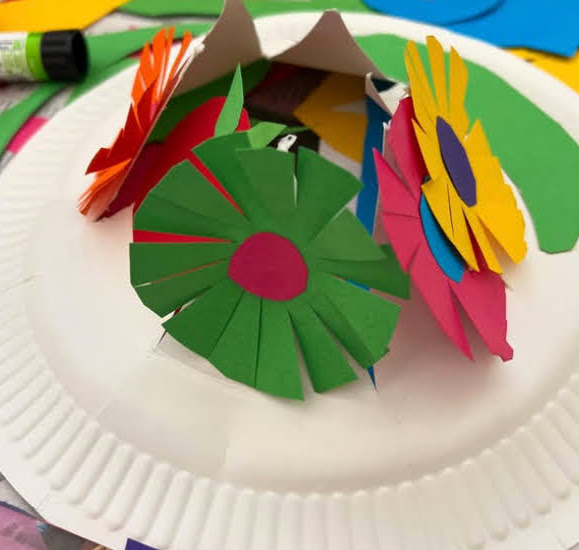 Halloween paper plate hat with various multicoloured flowers. One is green petals with bright pink middle.
