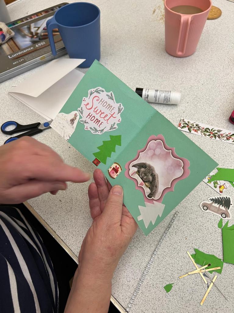 Christmas card made out of light blue/turqoise card. On one side of the card is a sticker saying home sweet home in red and white with a green christmas tree on the lower left side. On the other half of the card is a pink sticker and flowers and a white tree in the lower right corner.