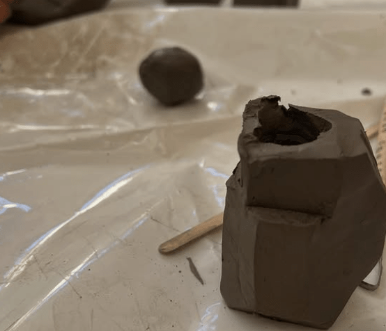 image of a pot with a middle and a ledge on its left side cut out of it. There is a tool in front of it and further in the center is a ball of clay.