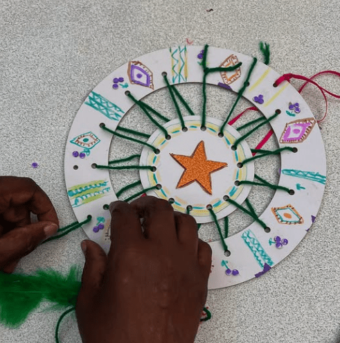 Dreamcatcher with an orange star in the middle. Its woven by green wool with a variety of pattern such as diamonds and vertical zigzag lines.
