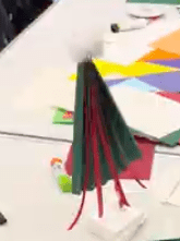 Photo of a tree thats halfway near completion. its is green inside it is red striped card. There are multiple coloured paper people are using behind it.