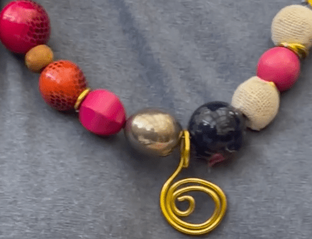 close up Photo of finished necklack. Has red, magenta, silver and black beads. In the middle is a curled up bit of gold wire as decoration.