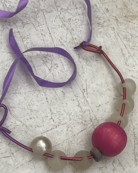 Finished necklace made of pink wire. It has white beads on it with a giant pink one in the middle. it's connected by a purple ribbon.