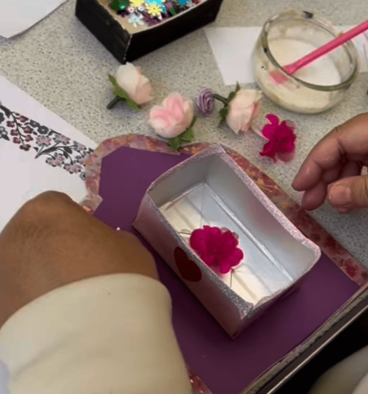 Shrine heart box 1: Has a purple base with a pink floral border. The box on top is metalic with a pink flower inside. on upper right is some other pink and purple flowers and glue.