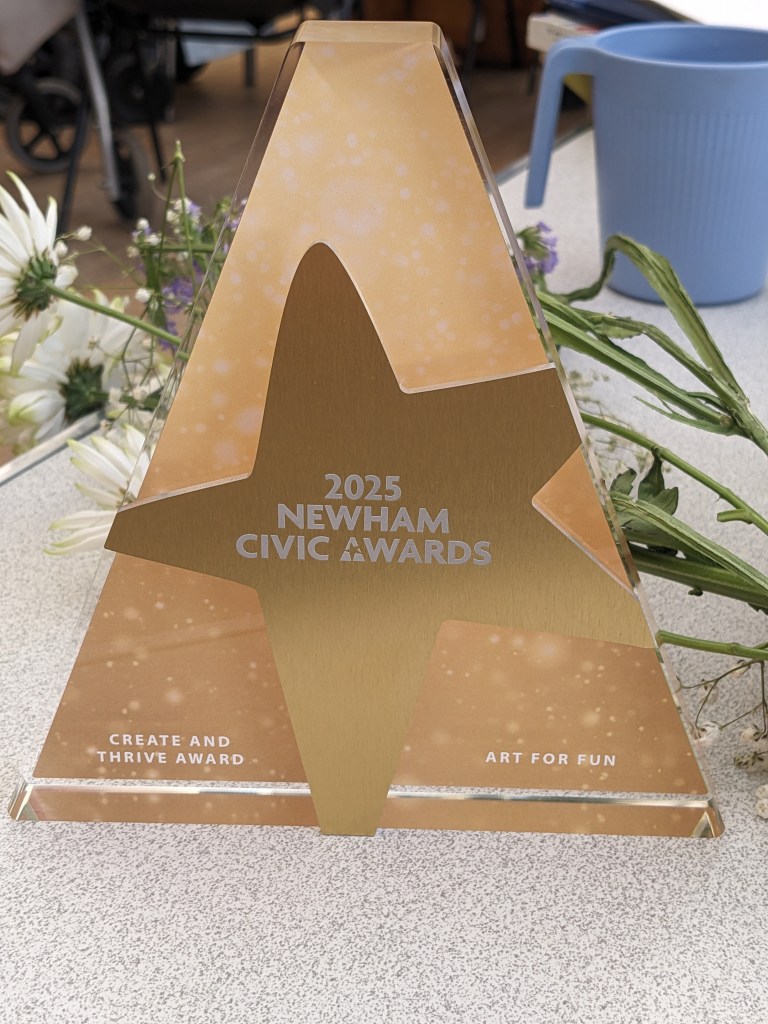 Photo of the Create and Thrive award. it is triangle shaped and gold with a star in the middle with white text saying 2025 Newham Civic awards. 